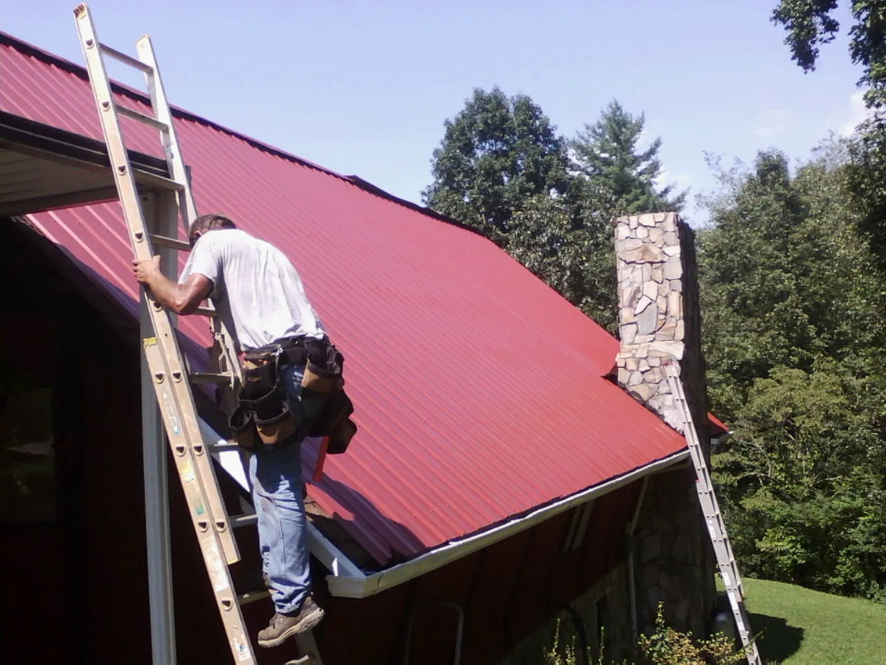 Roofer on a ladder beside a red metal roof and stone chimney — Soffit & Fascia Repair by Roofing Brothers in North Bend